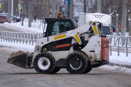 Small tractor loader machine clean and load snow and ice into a truck from a city streets after heavy snowfallの写真素材