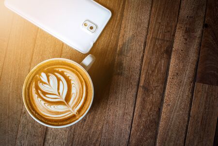 A white cup of coffee latte or cappuccino art and smartphone on wood tableの写真素材