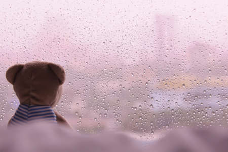 Lonely Teddy Bear sitting on bed and looking out at the window in rainy day. This photo has space background for text - vintage tone.の写真素材