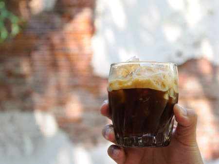 The hand holding a glass of Shaken Espresso mixing ice cube with coffee for cold drink to enjoy hot weather of the summer with the background of old brick wall which have space for text and backgroundの写真素材