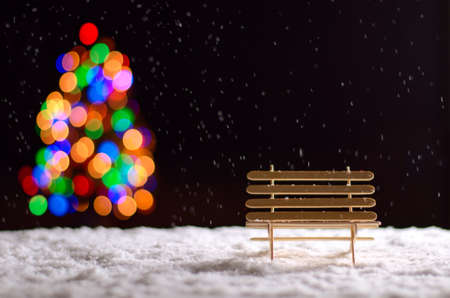 Wooden bench when snowing on the floor in winter season and colorful bokeh lights of Christmas tree.