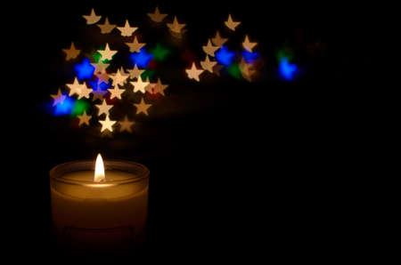 White aromatic glass candle with flame and colorful star shape bokeh on dark background for Christmas holiday.の写真素材