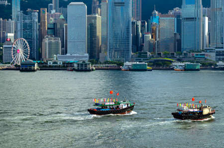 The Local ferry with China and Hong Kong flags sailing in front of beautiful skyline of Hong Kong city from Tsim Sha Tsui area face to Central area with many high buildings and sea view.のeditorial素材