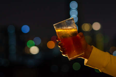 Hand with glass of beer toasting for celebration and party concept isolated on dark night background with colorful city bokeh lights on rooftop bar.の写真素材