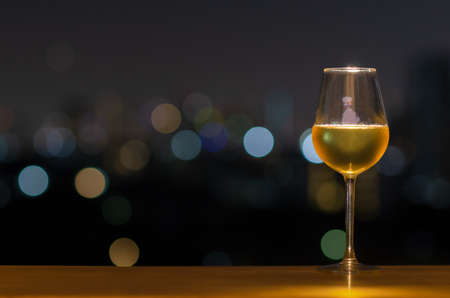 A glass of white wine on wooden table of rooftop bar with colorful bokeh of city light and space for text.の写真素材