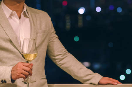 Businessman wearing grey color suit stands at the rooftop bar holding a glass of white wine with dark background of city bokeh lights.の写真素材