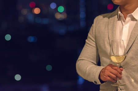 Businessman wearing grey color suit stands at the rooftop bar holding a glass of white wine with dark background of city bokeh lights.の写真素材