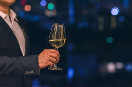 Businessman wearing navy blue color suit stands at the rooftop bar toasting a glass of white wine with dark background of city bokeh lights.の写真素材