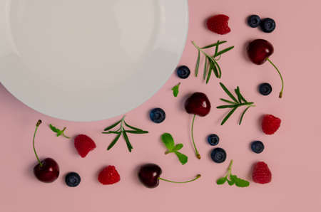 Fresh cherries, blueberries, raspberries, mint and rosemary leaf on top view with white plate and pastel pink color background for healthy food concept.の写真素材