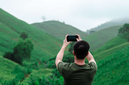 A male person using mobile phone taking a photo of greenery mountain with fog in the morning.の写真素材