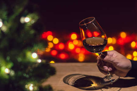 Hand toasting a glass of Rose wine on wooden table with Christmas tree and colorful bokeh light...