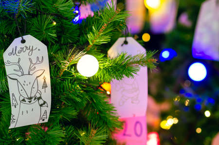 Paper hanging on Christmas tree for Holiday decoration with colorful lights on background.