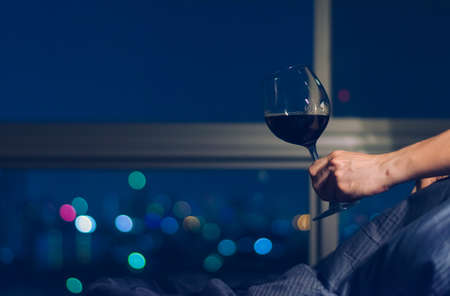A person holding glass of Red wine on bed that have colorful bokeh from city lights background.