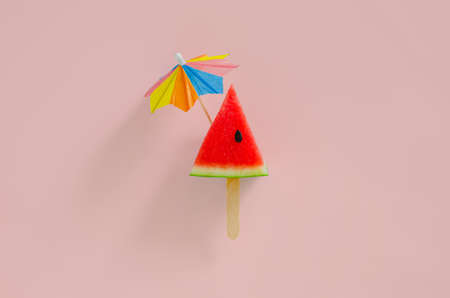 Red slice watermelon design as ice cream with stick that have cocktail umbrella on top on pink background. Minimal summer fruit concept.の写真素材