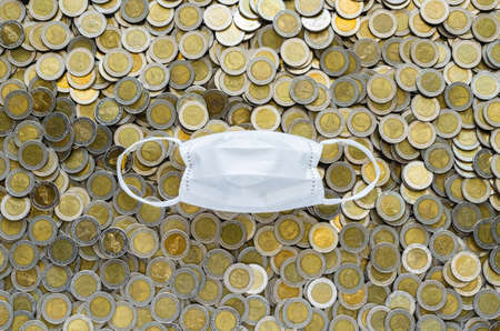 White face mask on background of Thai baht coins. Money saving concept in time of coronavirus pandemic.の写真素材