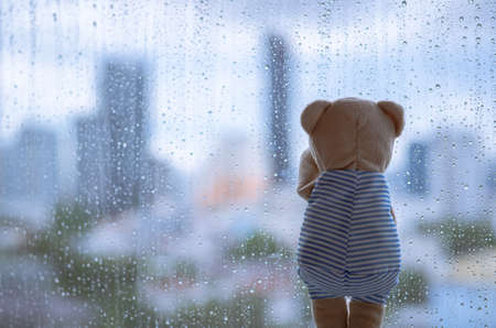 Teddy bear crying alone at window when raining with blurred city background.の写真素材
