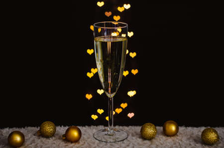 A glass of sparkling wine with golden christmas ornaments with love shape bokeh lights on dark background.の写真素材