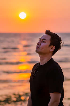 Asian man looking up to sky with sunset at the beach background. Positive mental health and emotion face expression.の写真素材