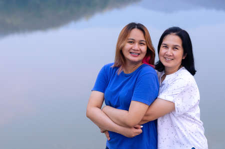 Single asian mother and her teenager daughter hugging together on lake background with shadow of...