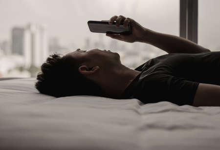 Asian man feels sad on bed looking and focus on smartphone. Internet social media addiction and mental health concept.の写真素材
