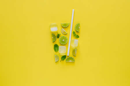 Mojito cocktail drink with lime, mint leaves, ice cubes and paper straw on yellow background. Minimal summer drink concept.の写真素材