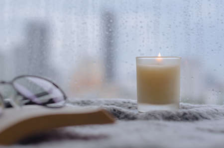 Burning aroma candle puts near by window that have rain drop in monsoon season with blurred city background. Zen and relax concept.の写真素材