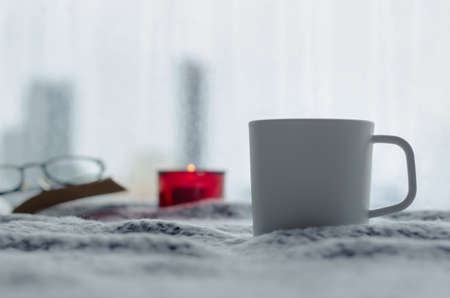 A cup of coffee puts on bed with aroma candle and book in winter season with blurred city background. Zen and relax concept.の写真素材