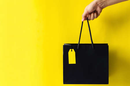 Hand holding black shopping bag with blank yellow price tag with yellow background for Black Friday shopping sale concept.の写真素材