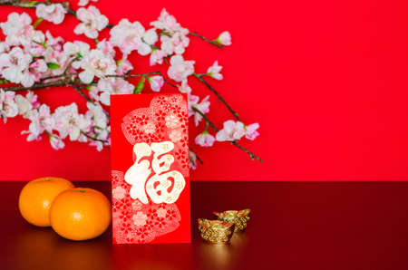 Red envelope packet or ang bao (word mean wealth) puts with oranges and ingots with peach blossom on red glitter paper background.の写真素材