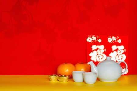 Chinese tea set put with ingots, oranges and red envelope packets or ang bao (word mean wealth) with shadow of peach blossom on yellow and red glitter paper background.の写真素材