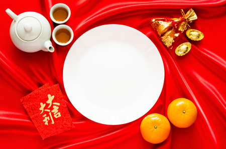 White plate on red satin cloth background with tea set, ingots, red bag (word means wealth), oranges and red envelope packets or ang bao(word means auspice) for Chinese new year concept.の写真素材