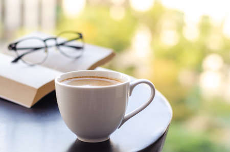 Selective focus of a white cup coffee puts on table at balcony with book and spectacles. Relaxing at home concept.の写真素材