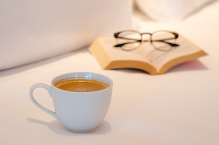 Selective focus of a white cup coffee puts on bed with book and spectacles in bedroom. Relaxing at home concept.の写真素材