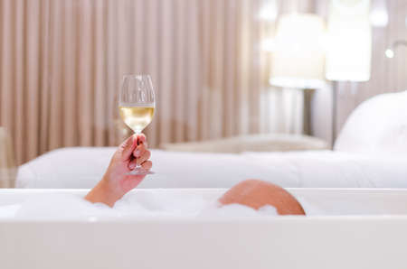A person holding a glass of white wine in bathtub at bed room. Relaxing at home concept.の写真素材