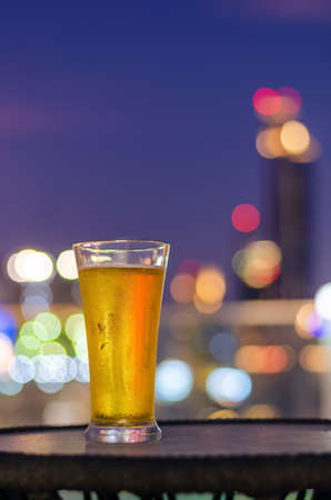 A glass of beer puts on table at rooftop bar with colorful city bokeh light background.の写真素材