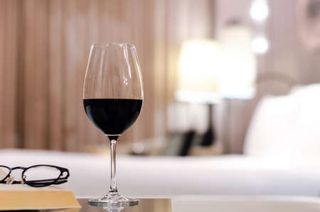 A glass of red wine with spectacles and book put on table in bed room. Relaxing at home concept.の写真素材