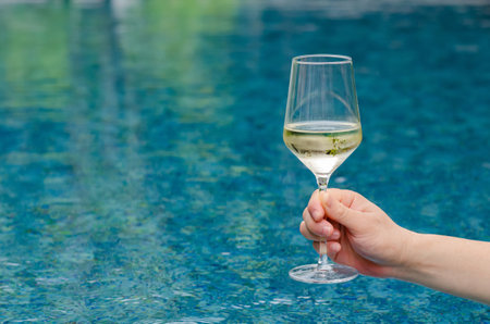 Hand holding a glass of white wine with swimming pool background. Holiday and summer drink concept.