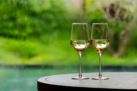 Two glasses of white wine on wooden table to celebrate for a couple with blurred swimming pool and garden background.の写真素材