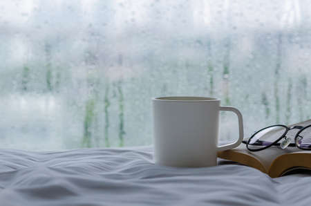 A cup of coffee with book and spectacles on bed in morning with rain drop on window. Stay home and relaxing concept.の写真素材