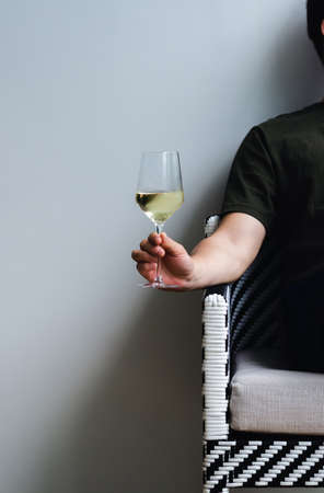 A glass of white wine holding by a person sitting on chair with shadow on white wall background.の写真素材