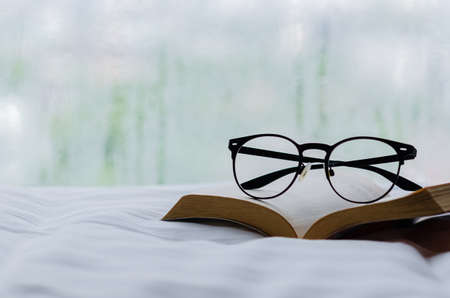Selective focus on spectacles and book put on bed in morning with rain drop on window. Stay home and relaxing concept.の写真素材