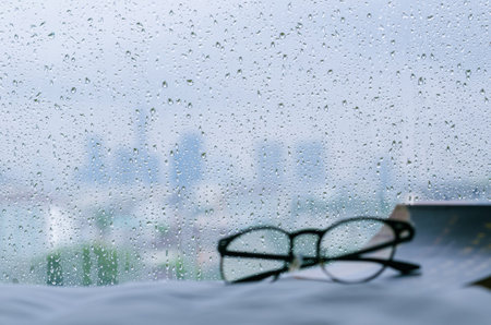 Rain drop on glass window at day time in monsoon season with blurred spectacles and book background. Stay home and relaxing concept.の写真素材