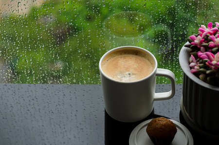 A cup of hot coffee with small cake on table in morning with rain drop on window and green background. Stay home and relaxing concept.の写真素材