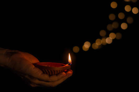 Hand holding the clay diya lamp lit on dark background with bokeh lights. Diwali festival concept.の写真素材