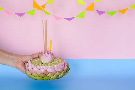 Pink lotus petal krathong that have 3 incense sticks and candle decorates with lotus and crown flower holding by hand for Thailand Loy Krathong festival on pink and blue background.の写真素材