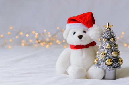 Selective focus on Santa claus teddy bear eyes who wearing hat sitting with blurred focus Christmas tree on white cloth background with lights.の写真素材