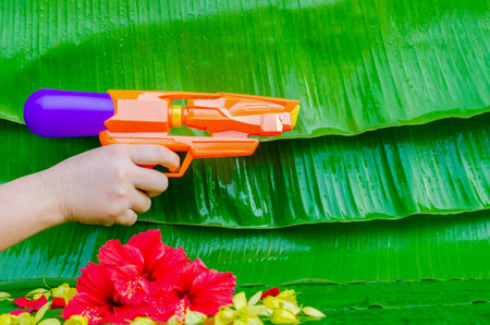 Hand holding water gun with colorful flowers on wet banana leaves background for Thailand Songkran...