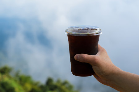 Hand holding a glass of iced coffee with blurred mountain and cloud background. Summer drink concept.の写真素材