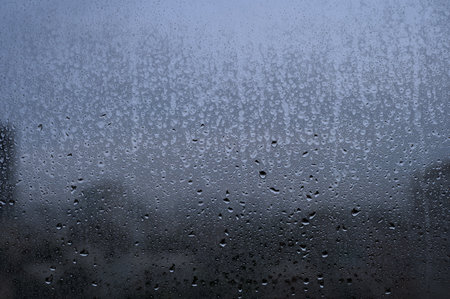 Raindrops on a glass window with a blurry cityscape in the background on a cloudy day.の写真素材