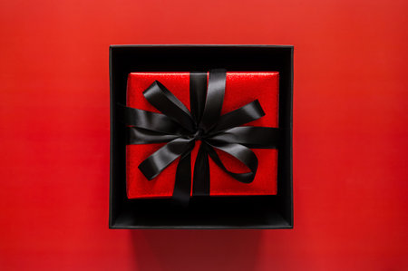 Black Friday concept with an open black box reveals a red gift with a black ribbon on a red background for signaling a secret deal.の写真素材